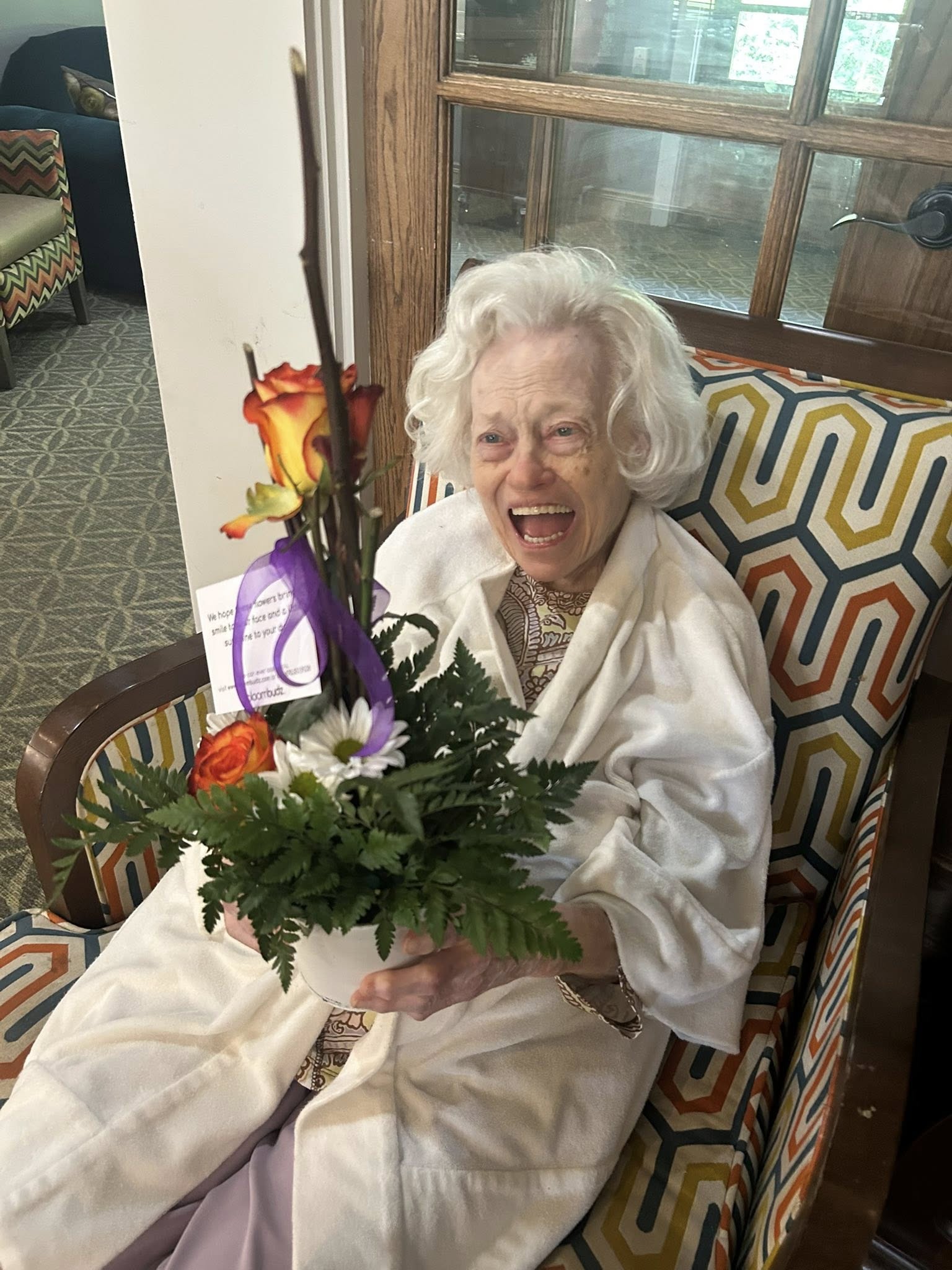 Woman in a white robe holding flowers in a room with a colorful chair and decorative elements.