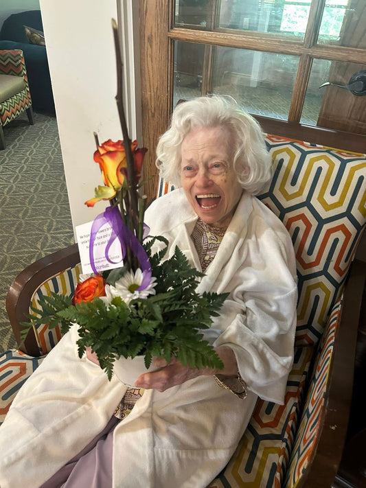 Woman in a white robe holding flowers in a room with a colorful chair and decorative elements.