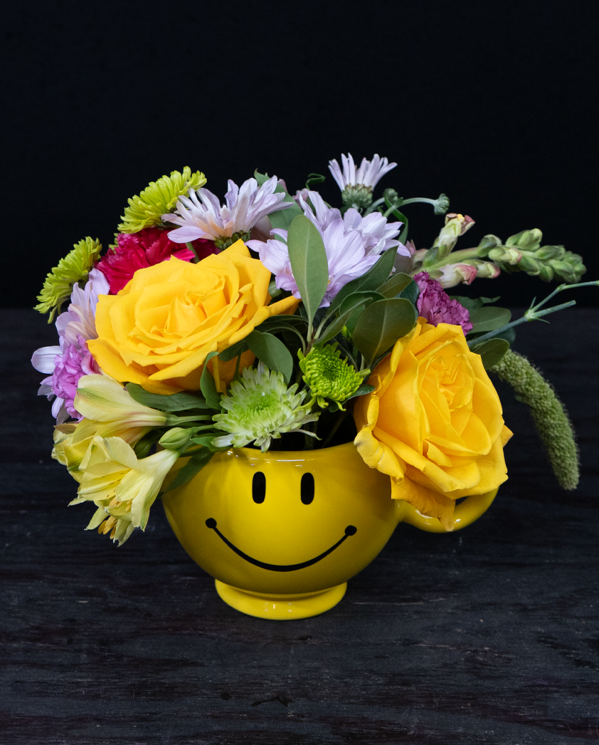 Pure Happiness yellow and lavender flower arrangement in a cheerful smiley-face mug